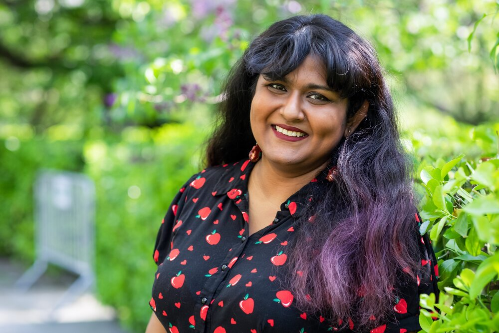 This is a photo of a Bengali femme with deep red lipstick, immaculate bangs, and long dark hair. She is smiling and wearing a black button-up shirt with red apples and small hearts on it. She’s in front of a verdant, leafy backdrop. The subject of the photo is Nibedita Sen, and you can find the link to her website in the introductory paragraph just below this photo.