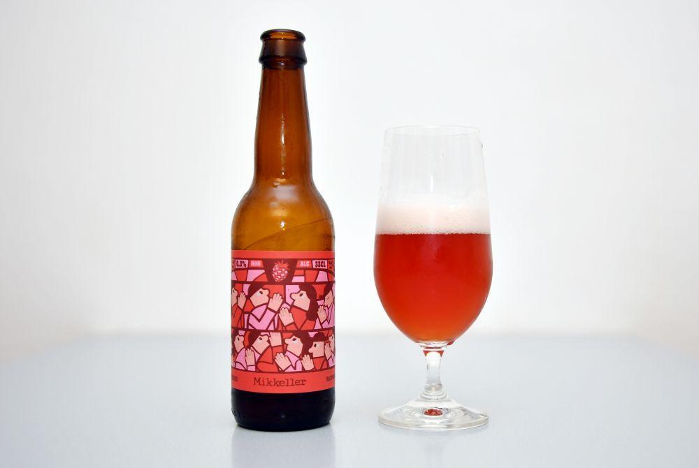 A glass of Mikkeller Limbo Raspberry beer sitting on a table beside beer bottle