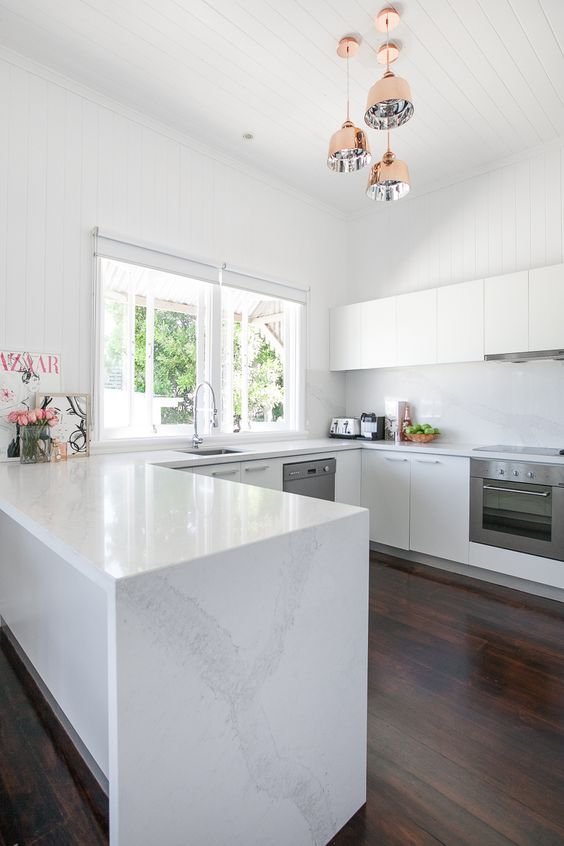 white u-shaped kitchen with flat panel cabinets, dark wood flooring, hanging brass pendant lights and a peninsula with white marble countertops