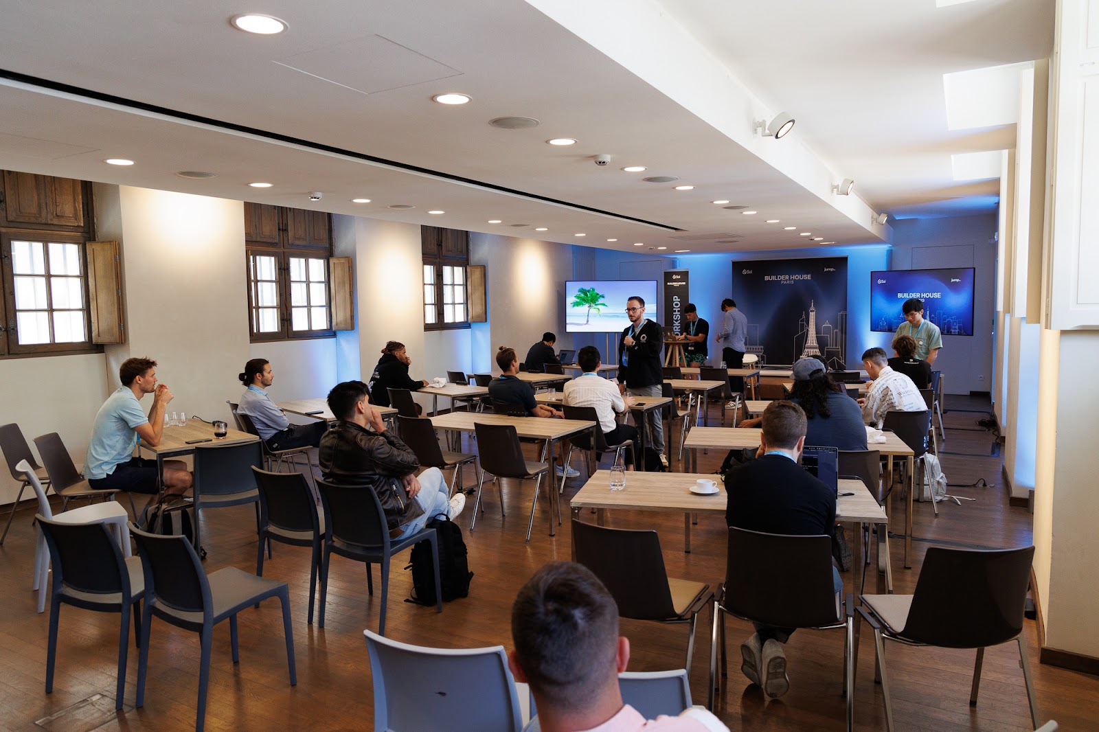 a room with people at desks listening to a speaker