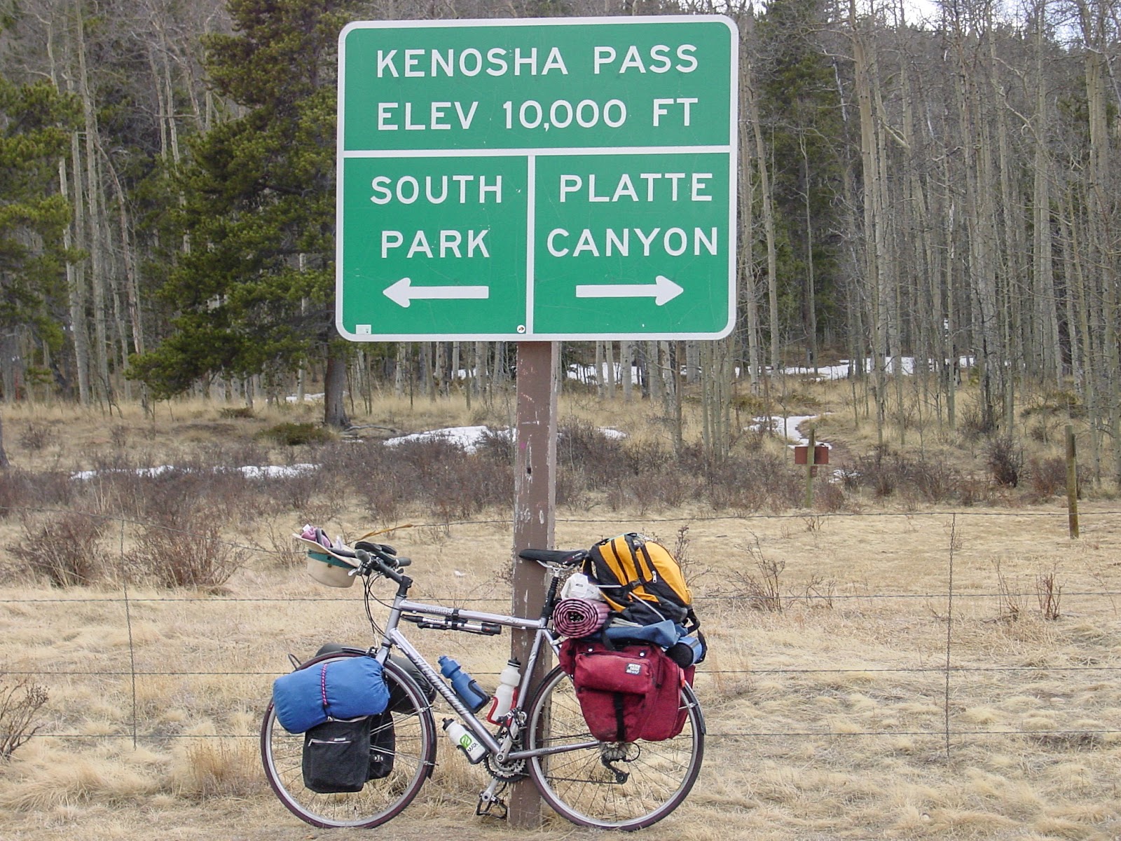 Bike leans on a sign that says: "Kenosha Pass Elev 10,000 ft." Pointing to the left it says: "South Park" and to the right it says: "Platte Canyon"