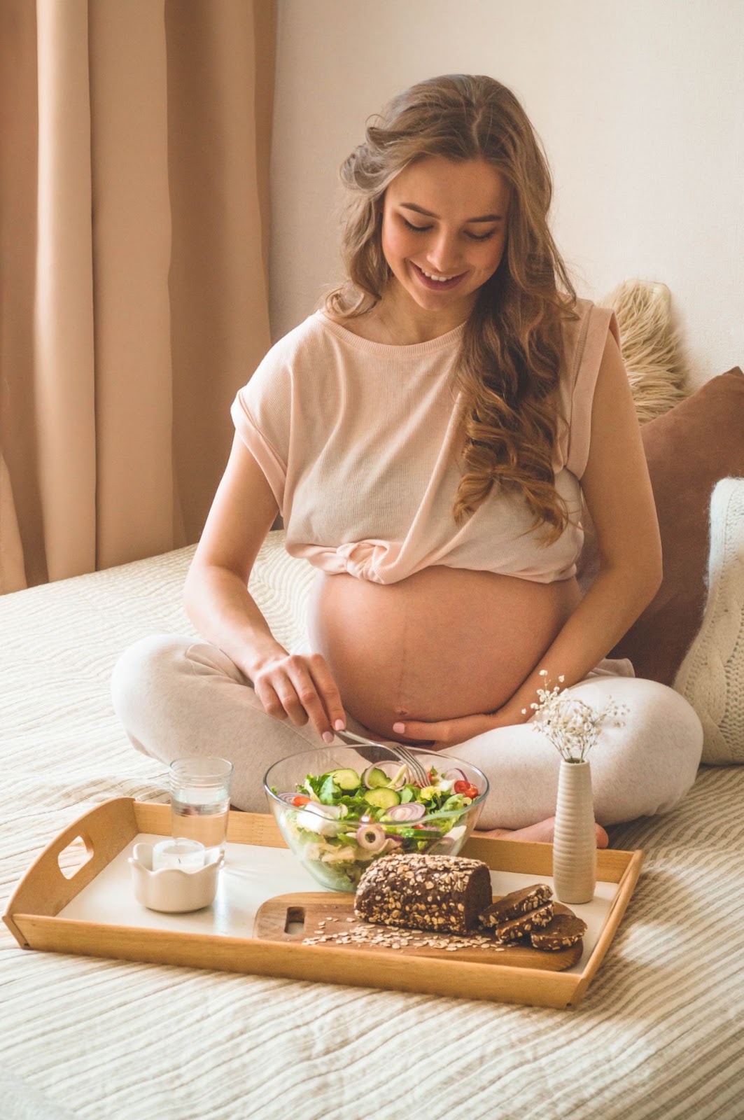 Mulher grávida, loira de cabelos longos e ondulados. Ela está usando uma calça de moletom cinza claro, com uma blusa rosa claro curta aparecendo a barriga. Ela está sentada na cama, com um café da manhã nutritivo em sua frente. Há uma salada, pão e leite. Também há um mini vaso de flores junto do café da manhã, e atrás da menina há três almofadas. 