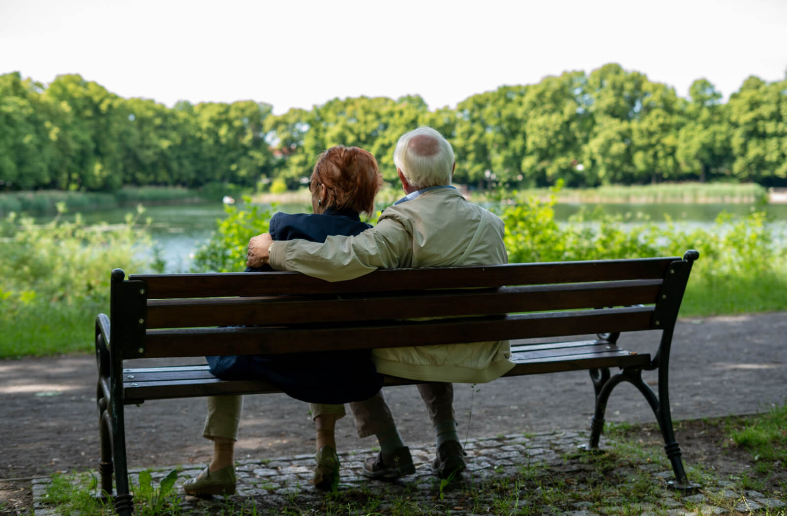 A female senior in a blue jacket and a male senior in a light brown jacket are sitting beside each other on a bench while watching the lake.