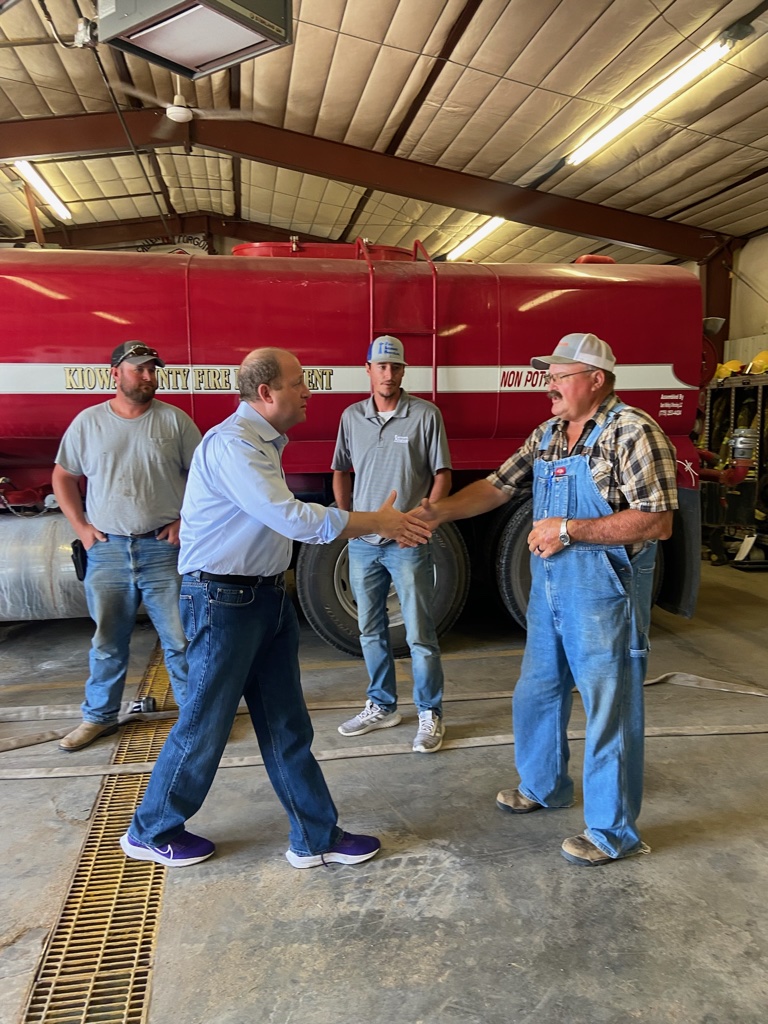 Governor Polis shaking hands with a man in overalls in a fire station.