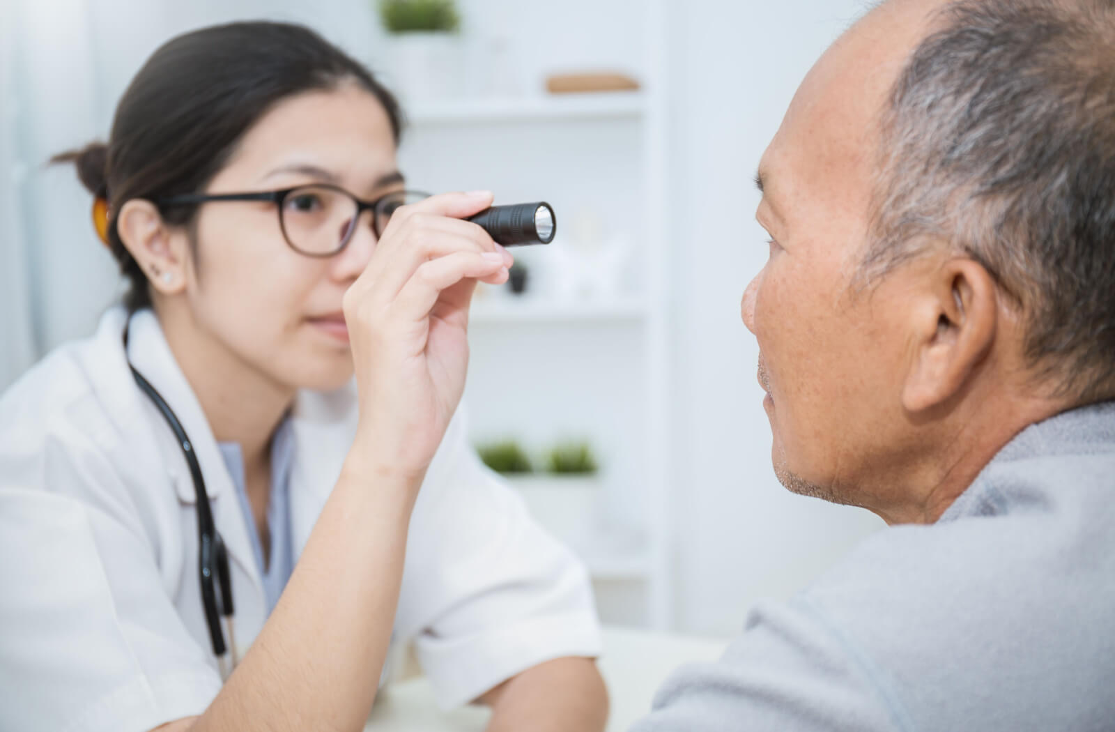 A senior man facing his optometrist as she shines a small flashlight into his eye.
