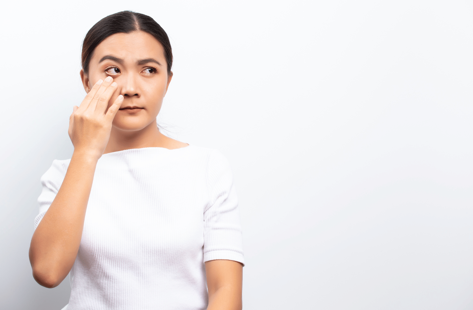 A woman standing against a white background, touching underneath her eye due to eye pain