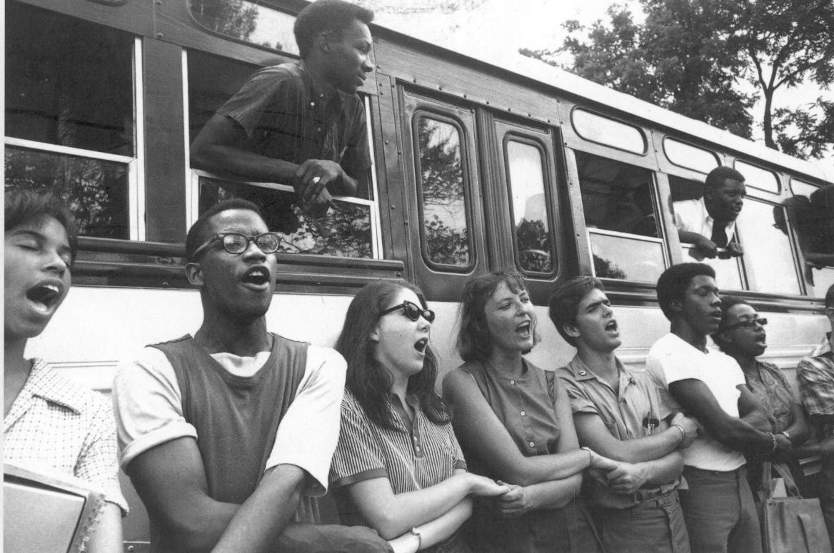 Freedom Summer volunteers holding hands and singing freedom songs beside a school bus before they prepare to travel to Mississippi. 