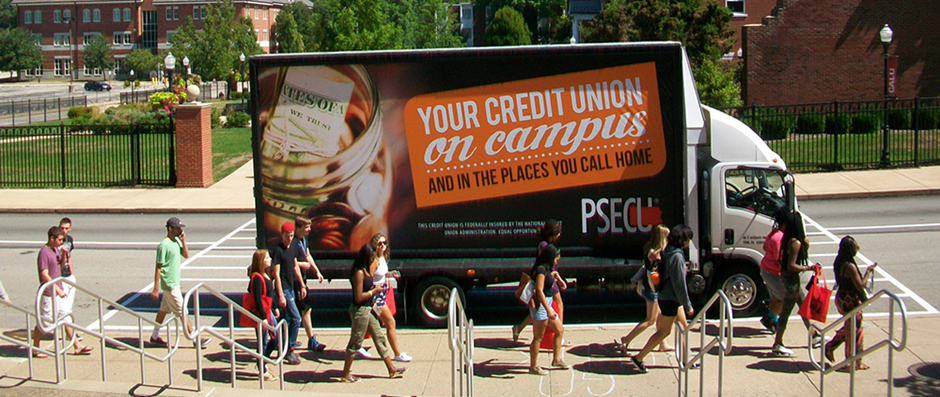 A parked truck on the roadside of a college campus, featuring an advertisement for the PSECU brand. The text on the side reads "YOUR CREDIT UNION on campus, and in the places you call home." The accompanying poster ad displays an image of a jar filled with coins, symbolizing savings and financial security.