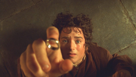 Elijah Wood in The Lord of the Rings. Frodo, a Hobbit with curly brown hair and bright blue eyes, is lying on his back on a grey stone floor. His hand is stretched out, reaching towards a golden ring above him.