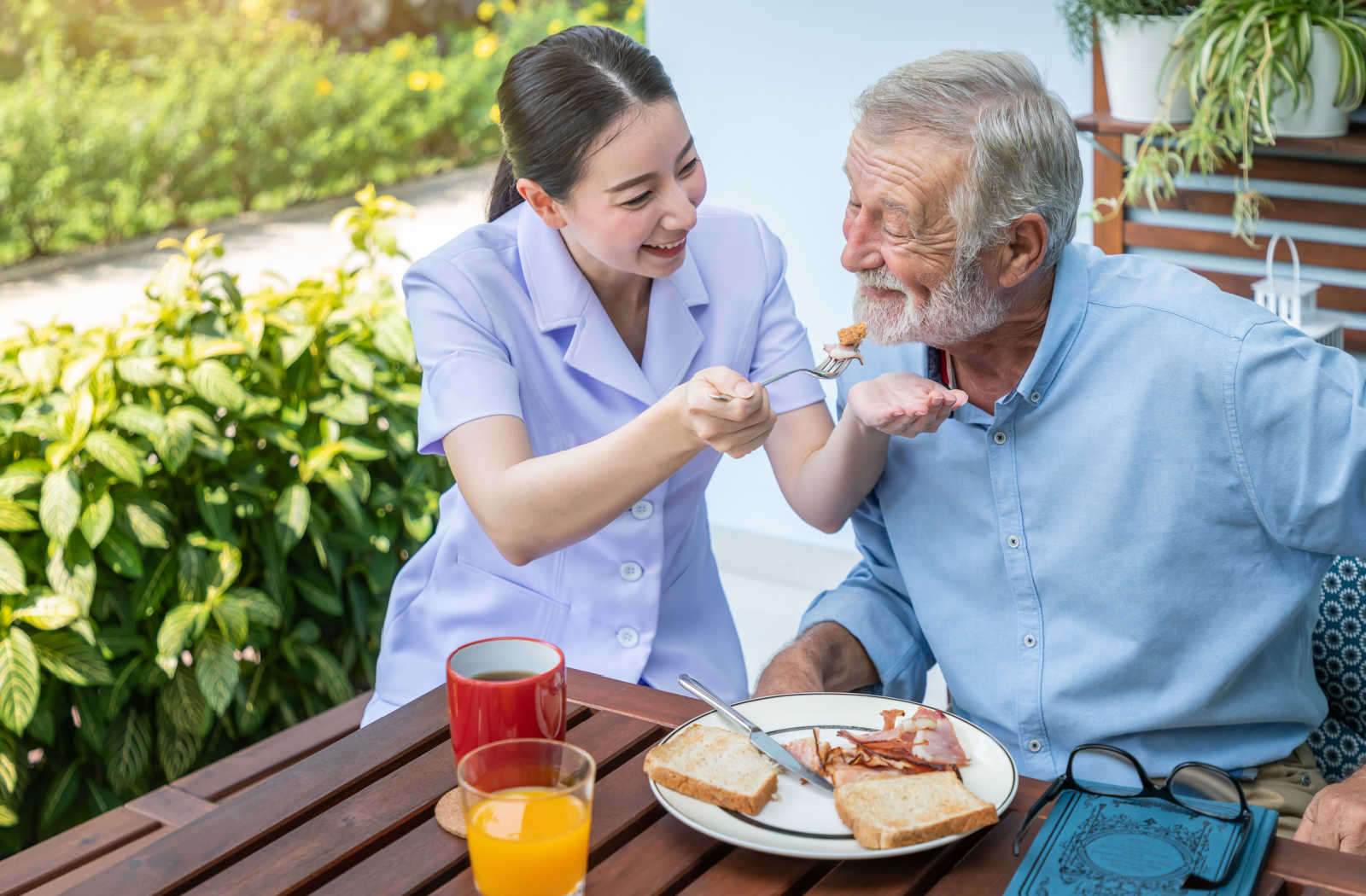 A memory care facility staff assisting a senior man to eat his breakfast. Both of them smiling.