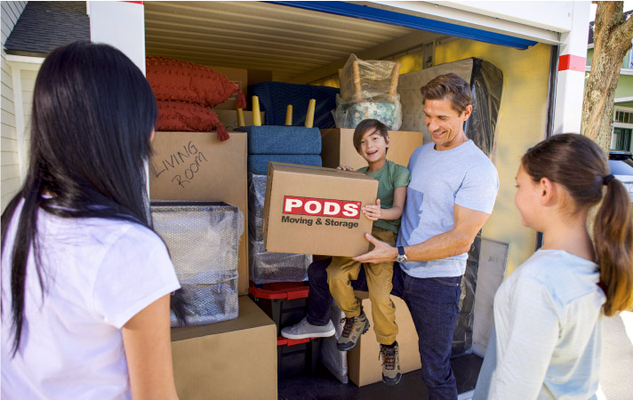 A family of four smiling together as the dad helps his young son load a moving box into their PODS portable moving container.