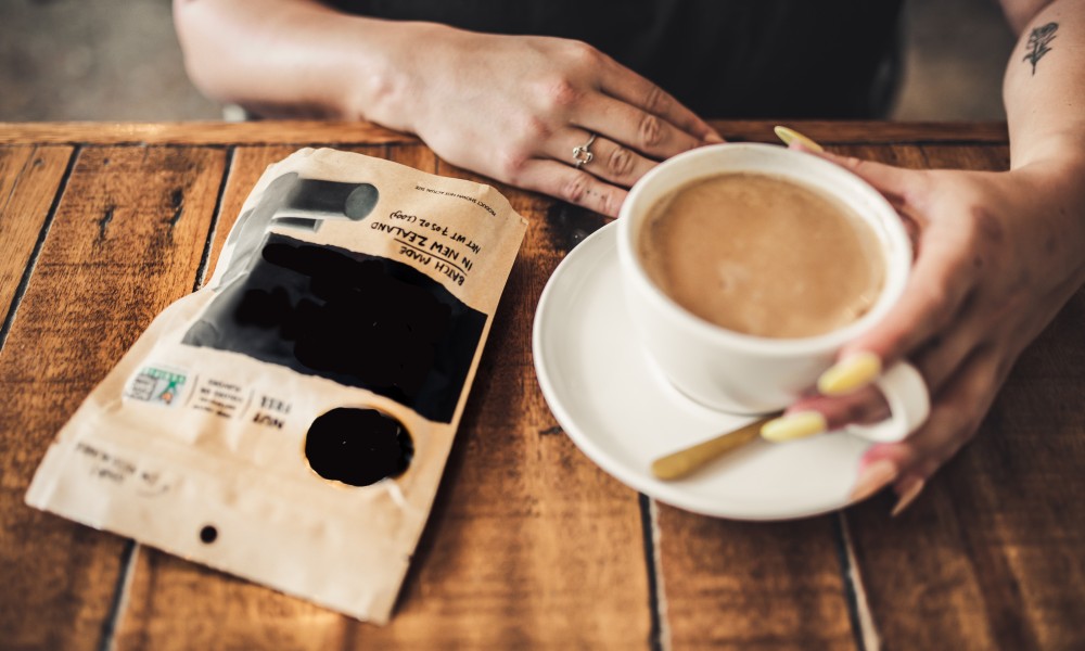 Image of customer with coffee in porcelain cup next to unbleached multilayer kraft paper coffee bag on wooden table. 
