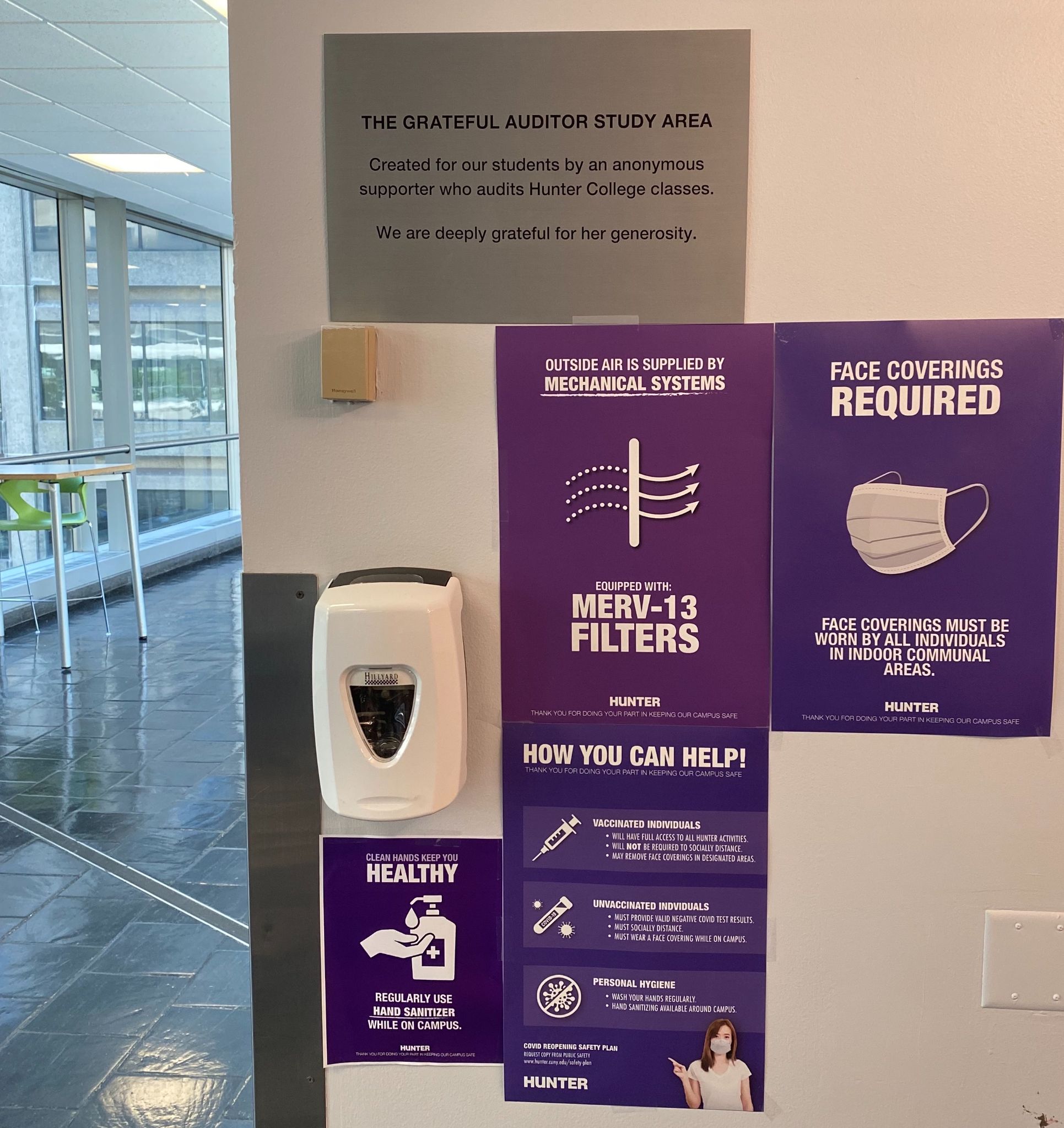 A wall in one of the halls at Hunter College is nearly covered in purple posters with Hunter's COVID-19 safety guidelines. One poster asks for all individuals to wear a mask indoors. Another poster displays distinct recommendations for unvaccinated people and unvaccinated people. A third poster encourages students to use hand sanitizer regularly, underneath a sanitizer dispenser. A fourth poster notes that mechanical ventilation systems are present in the hall. 