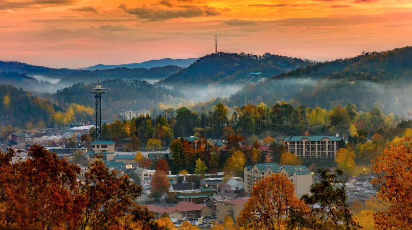 The sun rises over the city of Gatlinburg, Tennessee. Trees are turning hues of yellow and orange, and fog is moving in from between the mountains.