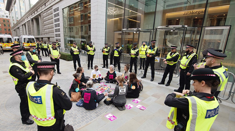 In front of a glass corporate building, a circle of 10 women rebels calmly hold hands. Stood around them are many police officers with arms crossed.
