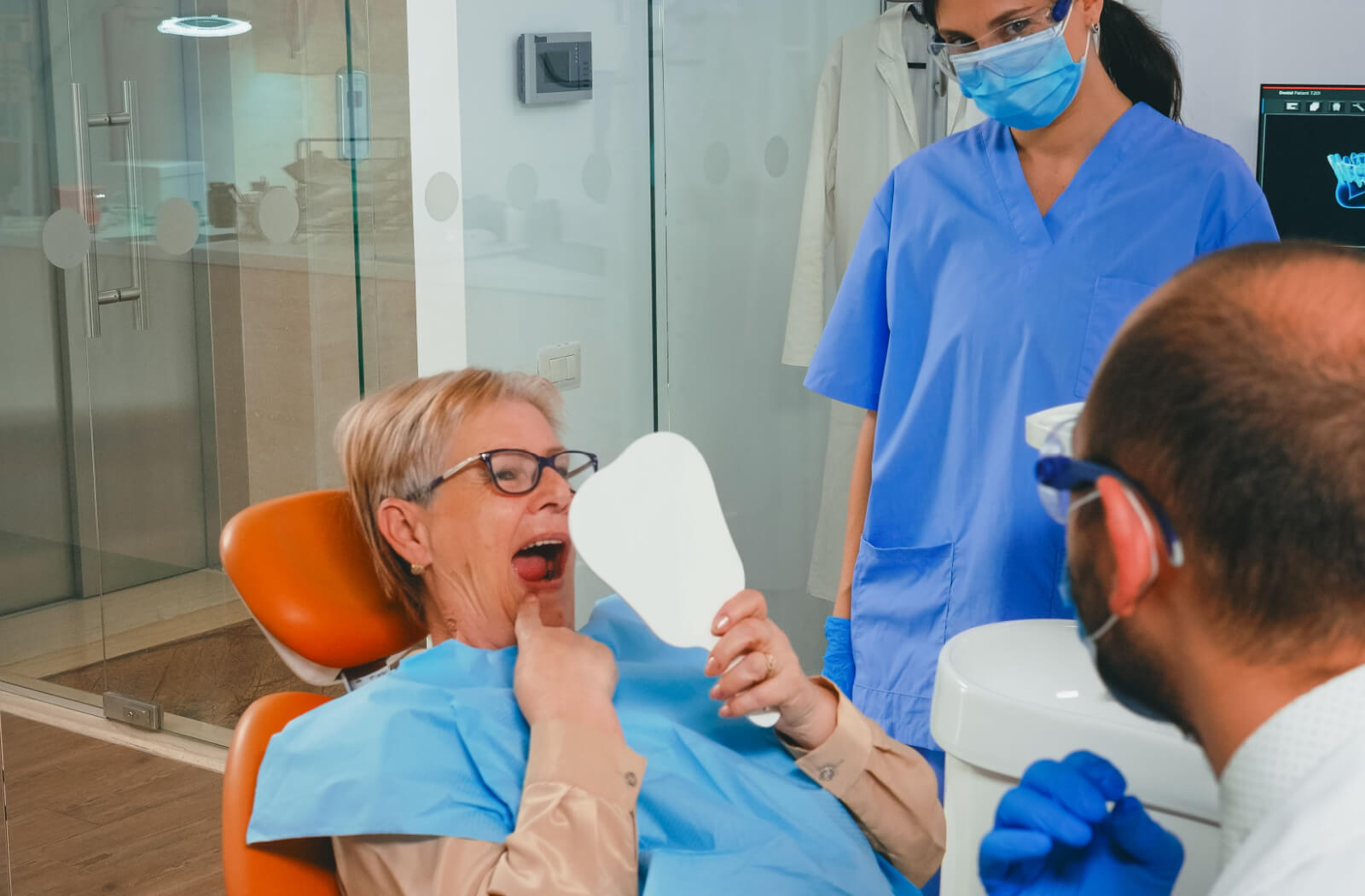 A senior woman sitting on a dental chair is looking at her teeth in the mirror after several minutes of dental cleaning.