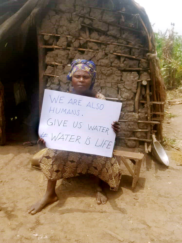 A congolese woman sits outside her mud hut with the sign 'We are also humans. Give us water. Water is life'.