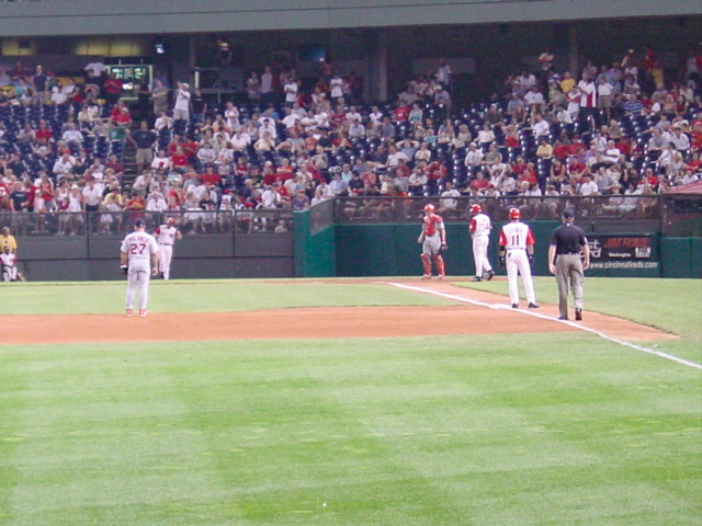 Between pitches the players look relaxed, standing still with a Reds player on third base. 