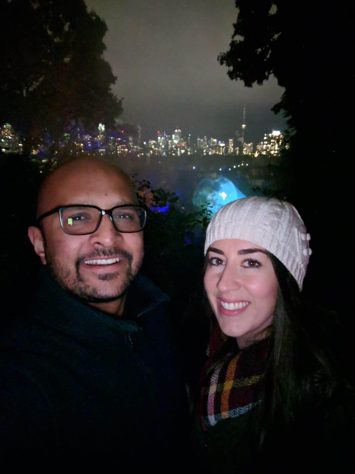 Melissa and her partner in coats smiling at the camera. She is also wearing a white toque and a scarf. They are in front of the Toronto city skyline at night.