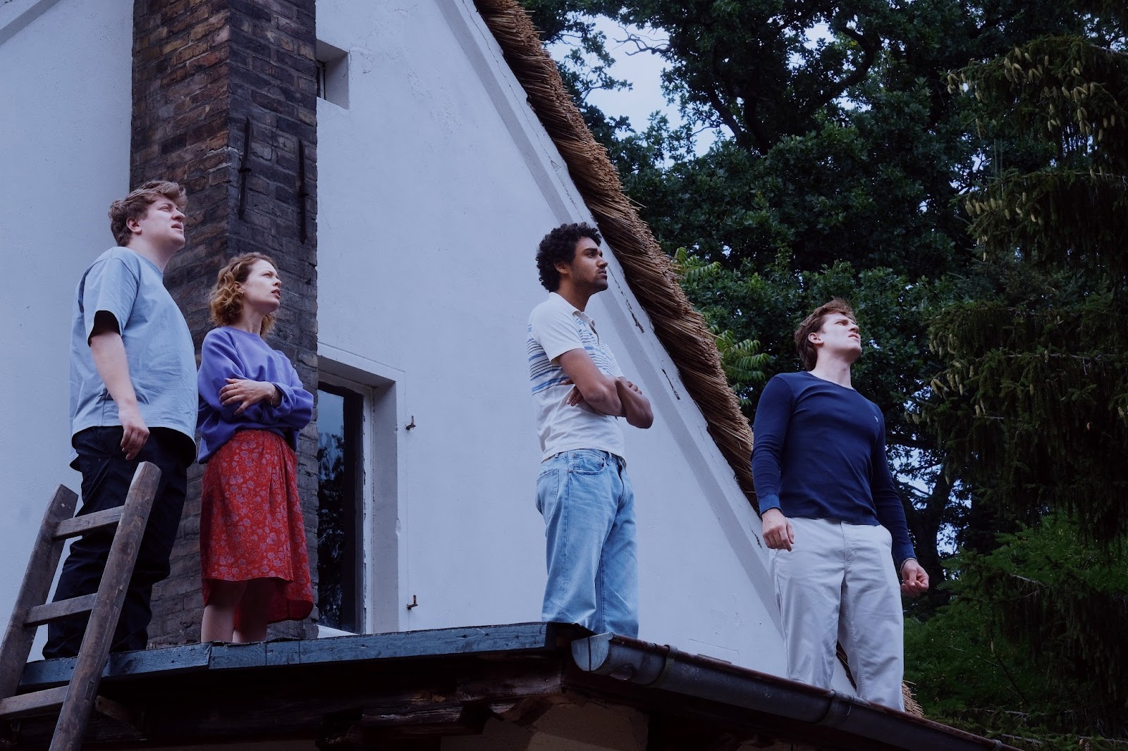 From the film Afire, four people stand on a roof of a house during the day, all looking up at the sky in the distance at something off screen.