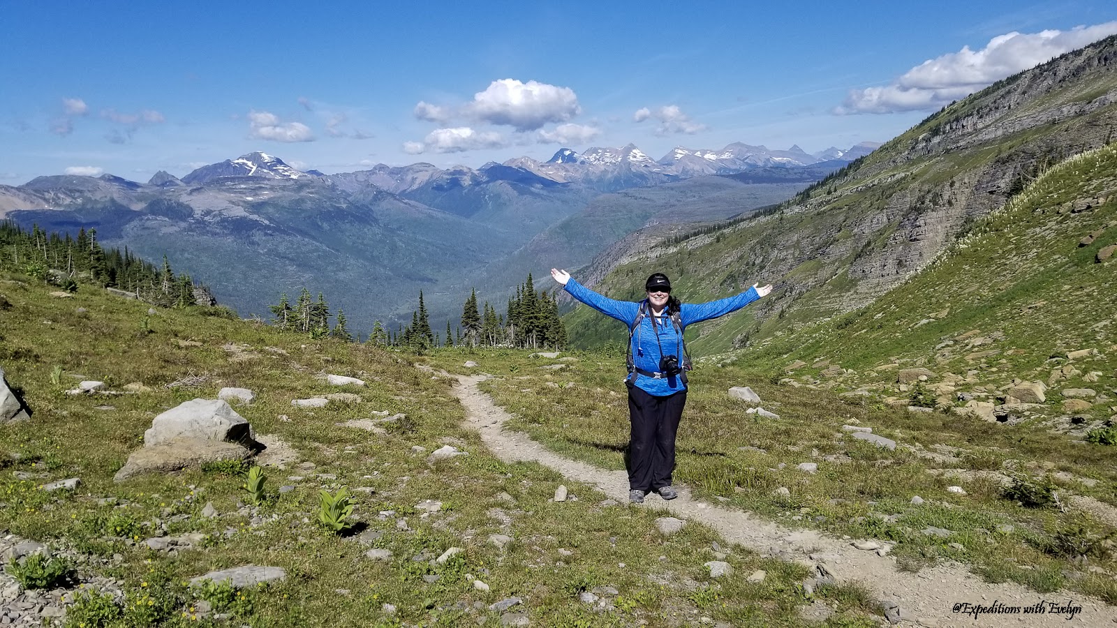 A female hiker embraces hiking in national parks with open arms on a mountain trail surrounded by small rocks, pine trees, and distance mountain peaks.
