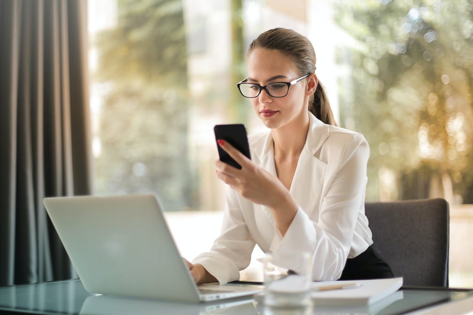 Woman wearing glasses checking her phone while working on her laptop