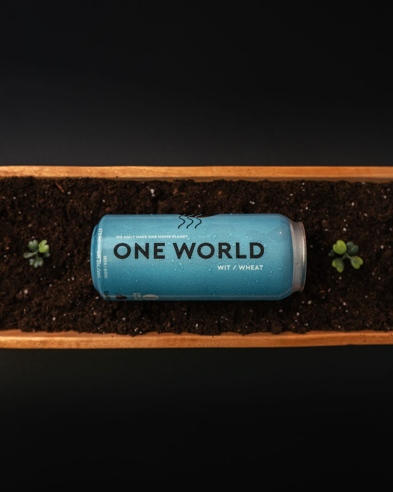 A Can of One World Wit laying on its side in a planter.  The can is turqouise and has the name of the beer down the side. 