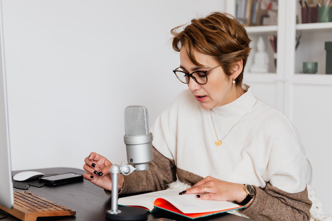 Free Female narrator in glasses reading out loud from book while sitting at desk with microphone and recording audiobook in cozy study Stock Photo