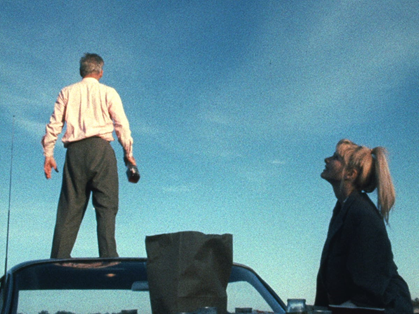 Michael Higgins and Barbara Loden in Wanda (1970). Mr Dennis (left), a middle-aged man with grey hair, wearing a white shirt and grey slacks, is standing on the roof of a car with his back to the camera, looking out at a clear blue sky. Beneath him to the right is Wanda, a young blonde woman with a high ponytail and a black coat, who is looking up at Mr Dennis and smiling.