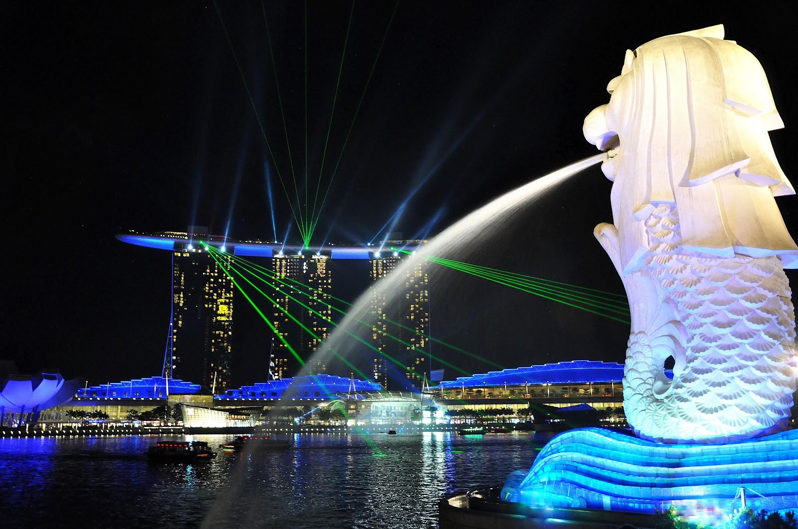 marina bay sands hotel illuminated by green and blue laser beams and white merlion statue in the foreground seen at night in singapore
