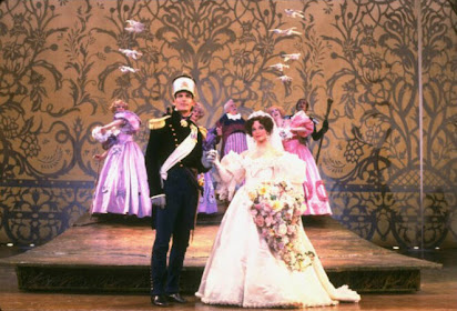 A stage with an ornately patterned backdrop. A woman and man stand at the front, arm in arm. They are dressed in wedding finery. There are two women behind them in pink dresses.