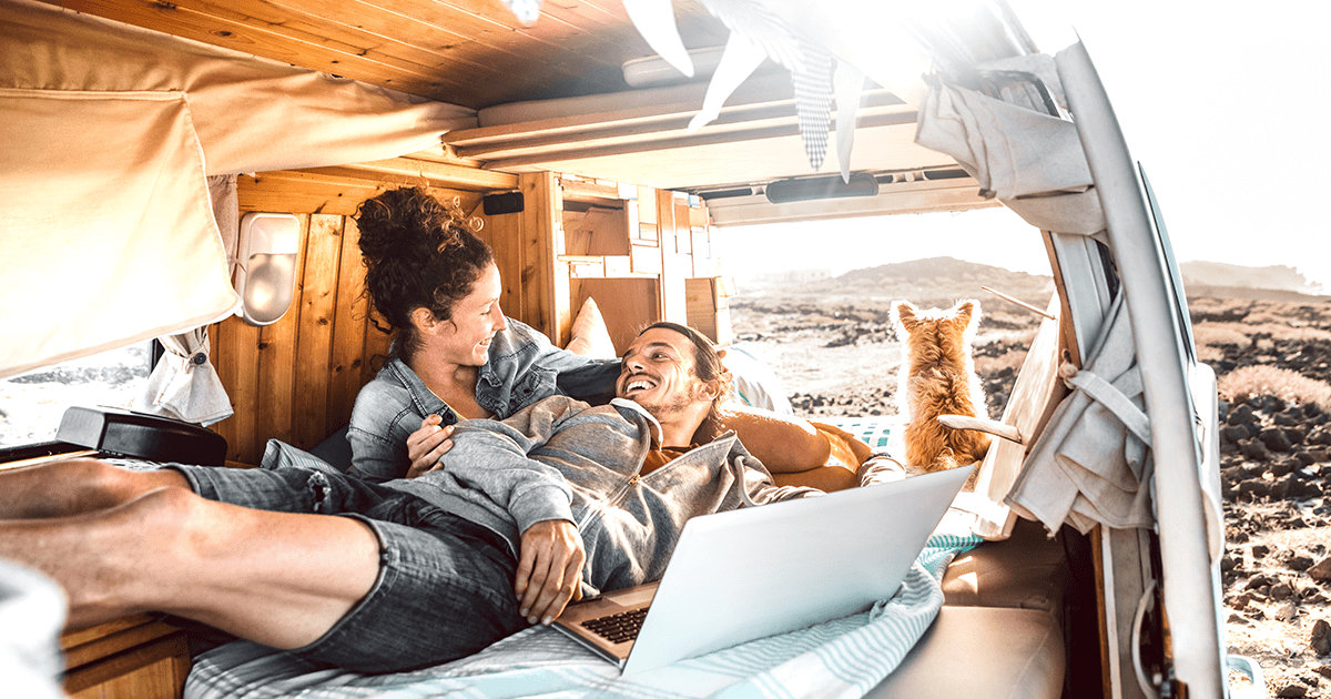 Man and woman laying in camper van smiling while terrier dog stares out at beach