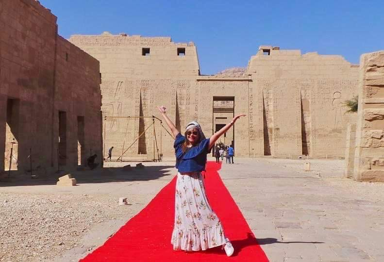 Image of Candy with her arms in the air, standing on a red carpet with an ancient Egyptian temple in the background