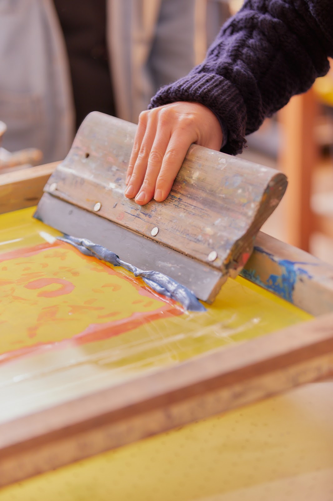 Image: A hand holding a squeegee with a wooden grip and metal inset, pressing into grayish blue ink on a yellow and orange screen in a wooden frame. Photo by Sarah Joyce.
