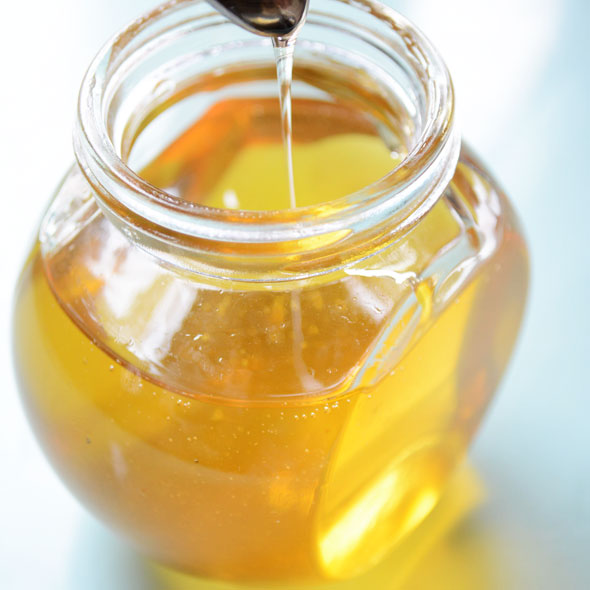 Photo shows a jar of golden honey, with a spoon raised above it and honey drizzling out of it