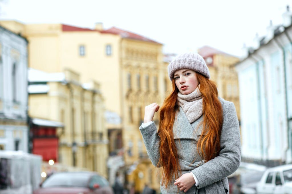 Street portrait of attractive redhead woman with long hair wearing warm winter wardrobe outfit posing at the street. 
