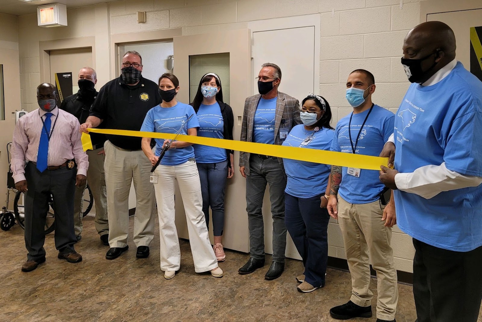 people wearing bright blue t shirts and holding a yellow ribbon and wearing face masks. One woman holds large scissors to mark the start of the jail's medication-assisted treatment program