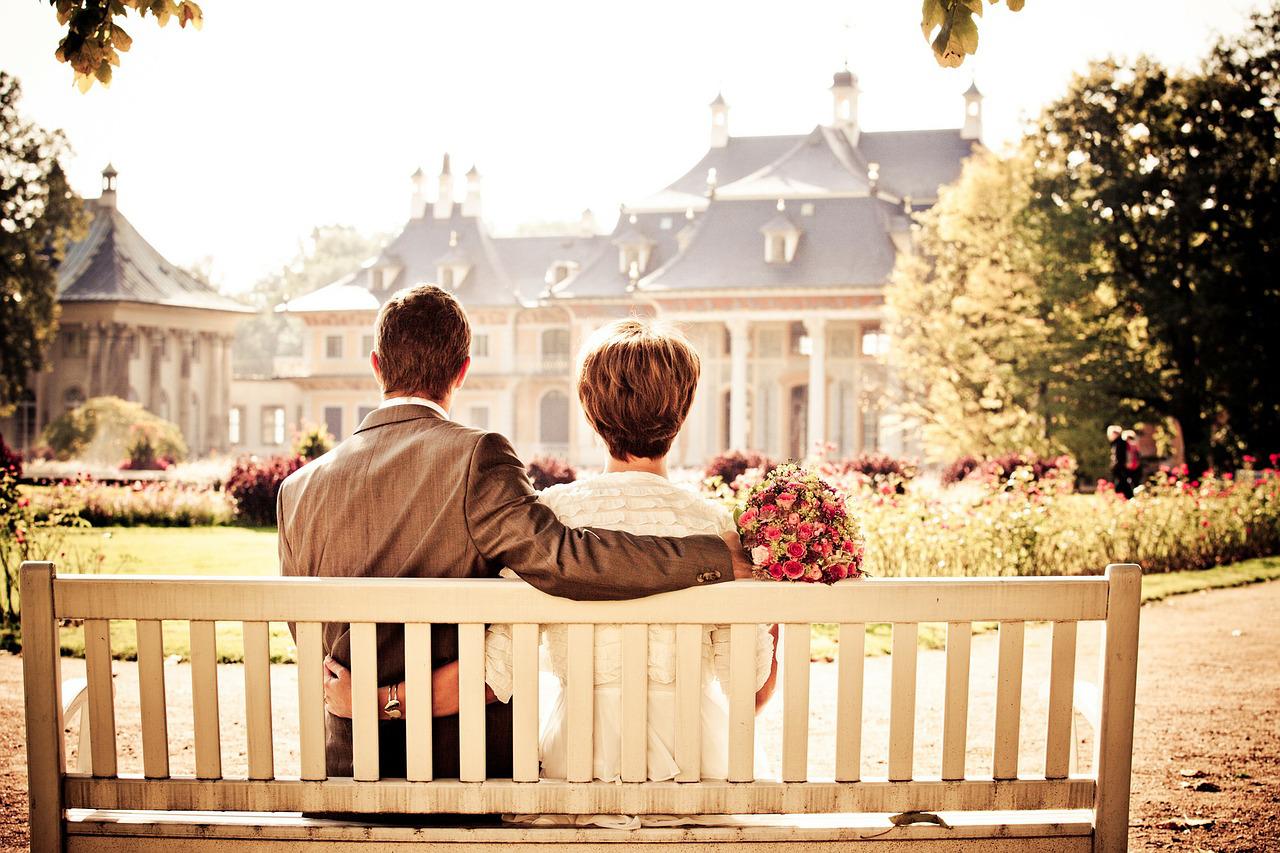 A man and woman sitting together on a bench looking at a mansion