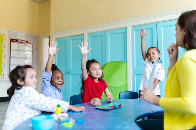 group of children sitting at a table raising their hands