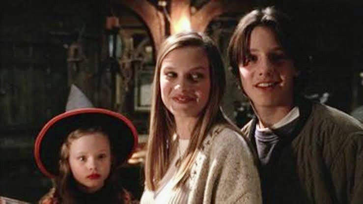 à esquerda, uma menina de batom vermelho e chapéu de bruxa preto. no centro, sorrindo, uma adolescente de cabelos loiros. E, à direita, um adolescente de cabelo escuro e blusa preta.