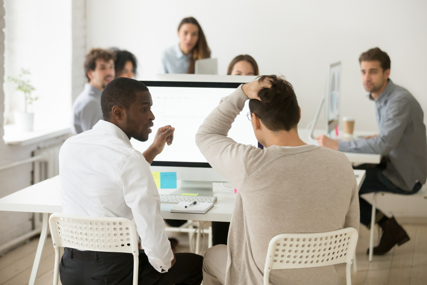 A imagem mostra em destaque algumas pessoas em torno de uma mesa de trabalho branco. No centro da foto, há um homem negro de costas, que olha para um homem branco que está ao seu lado, também de costas. Os dois parecem solucionar um problema no computador. 