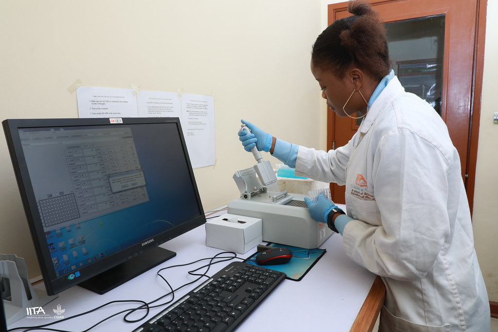 A young girl in a lab coat and latex gloves is dropping some type of liquid into a machine that is connected to a computer on the desk beside her.