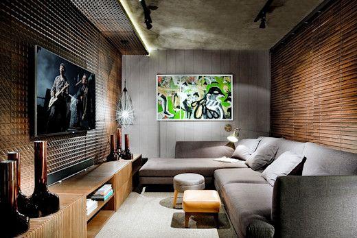 Sala com decoração escura, poucas luzes, revestimento de madeira na parede de fundo, sofá cinza, parede lateral com revestimento de madeira cinza e painel de TV de madeira com rack.