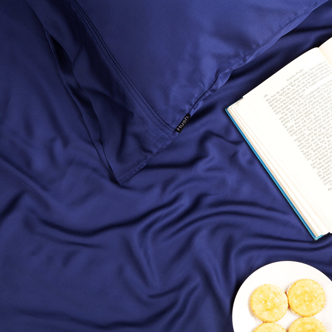 A plate of cookies and a book sitting on top of a set of Hush Iced 2.0 Bamboo sheets in Navy color.