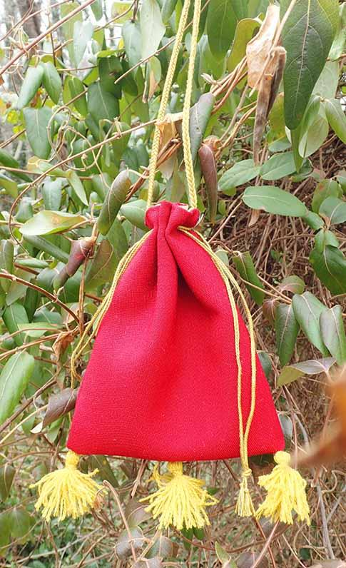 The completed red bag hanging off of a garden plant by its gold thread. Drawstrings are pulled on both sides and all three tassels hang at the bottom.