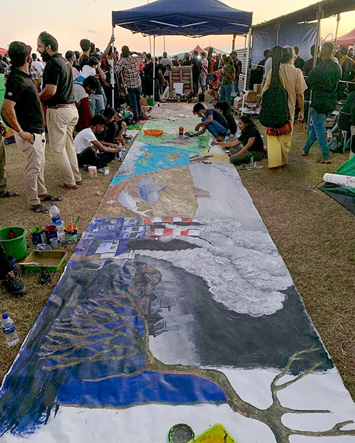 Protestors work on a very long banner laid on the ground, painting environment-themed scenes such as a tree growing from the ground, factory chimneys poluting the sky, creatures in the ocean