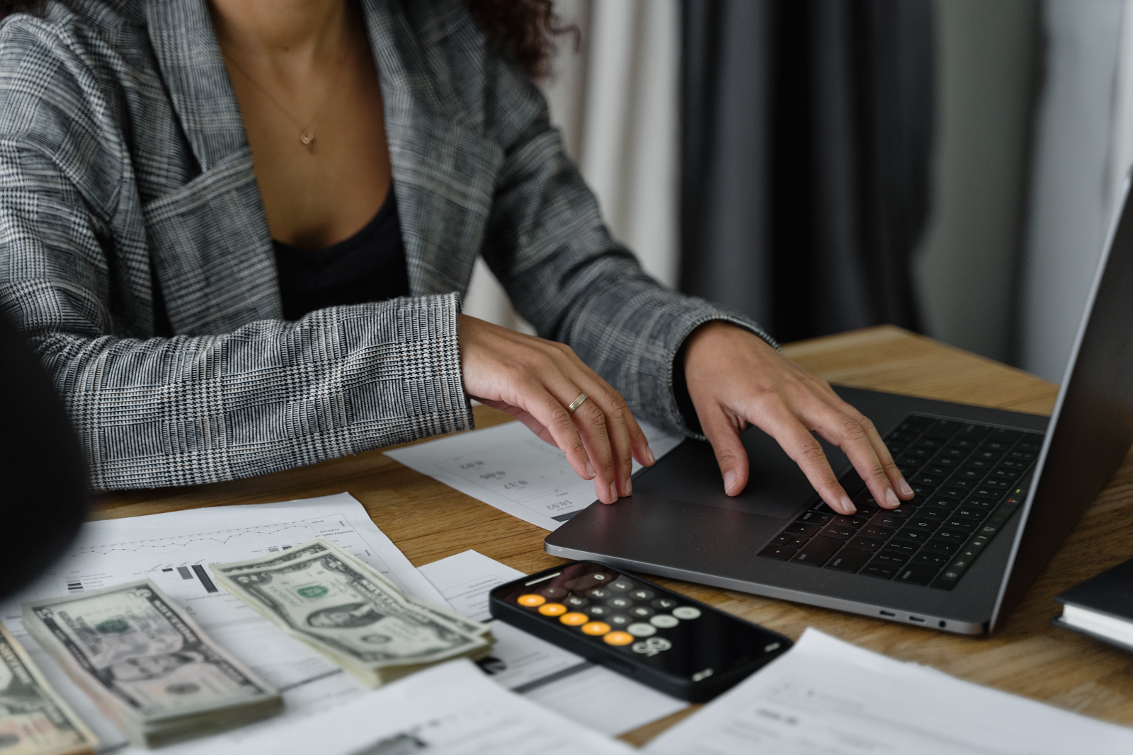 A person typing into a laptop. There are numerous papers and dollar bills on the table.