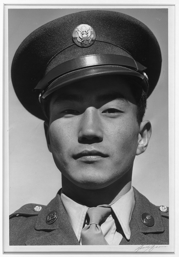 Portrait of a Japanese American soldier. He is looking at the camera with a slight smile, wearing a cap and uniform.