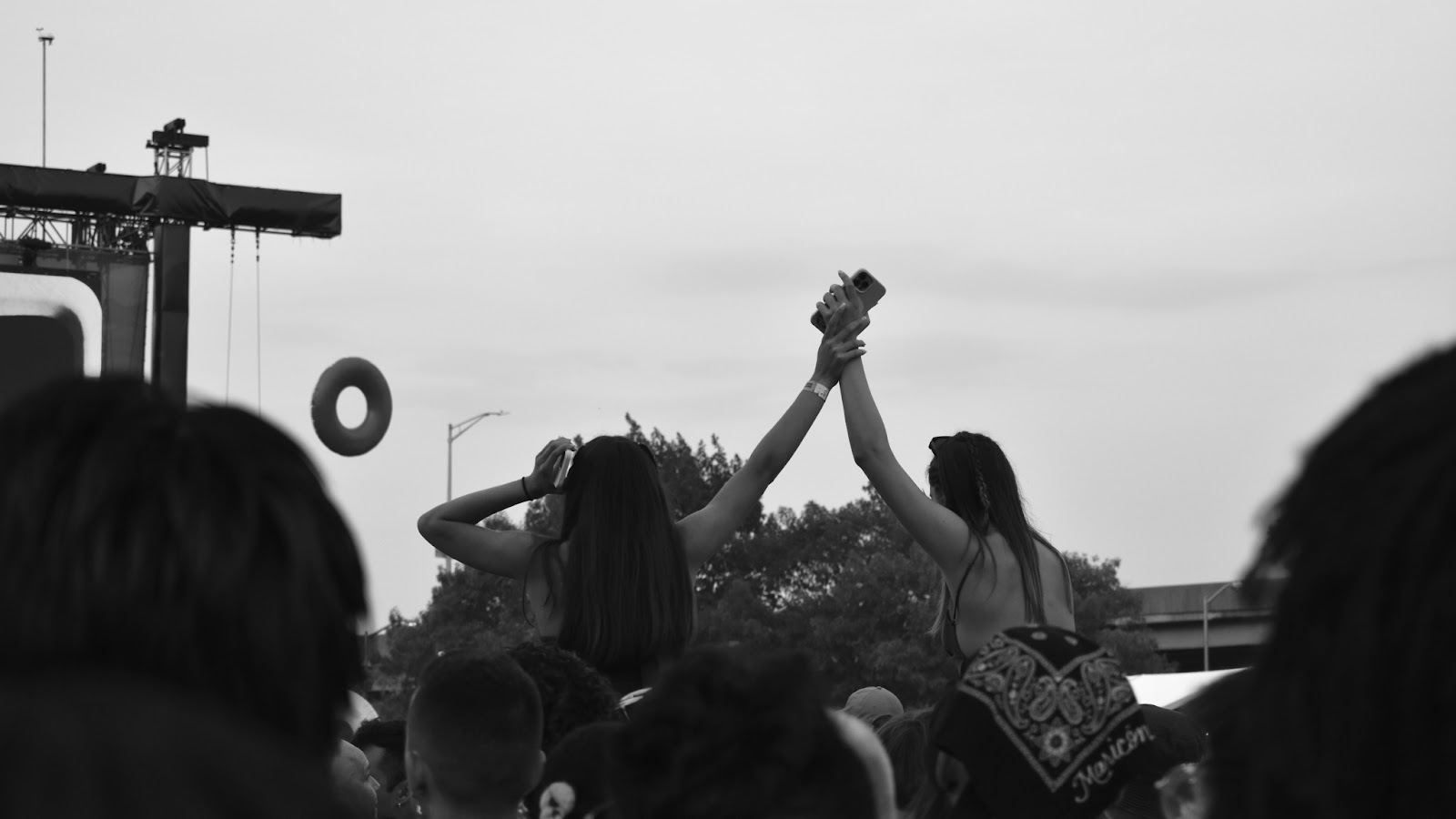 Two festival goers amidst a crowd hold hands raised above their heads whilst sitting atop friend’s shoulders. Their backs are turned to the camera as they face the main stage. The festival wristband is visible on one of their wrists.