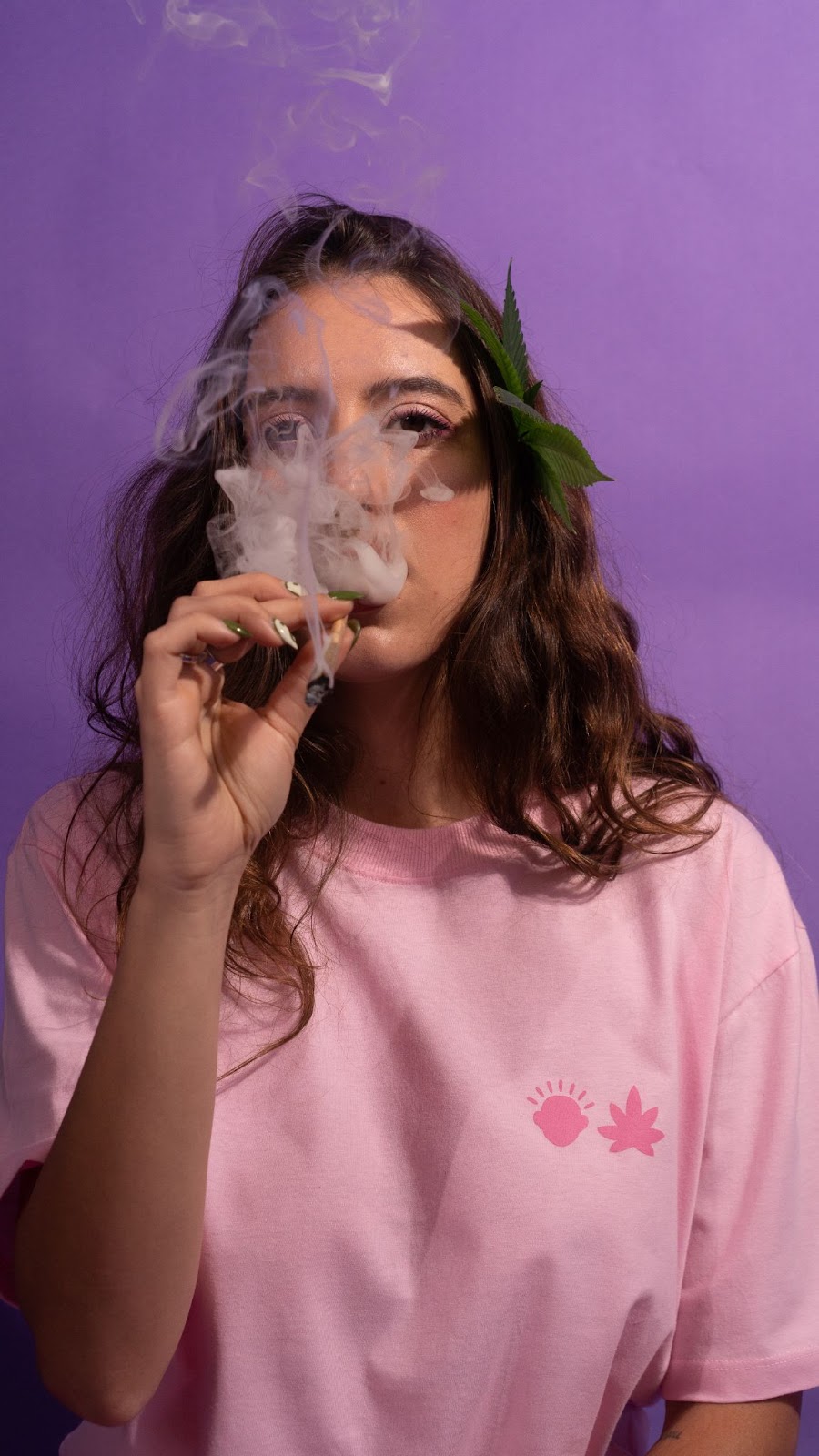 Foto colorida da Alice de camiseta rosa e folha de maconha no cabelo, fumando um com nossa piteira